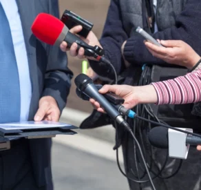 Group of journalists holding microphones, recorders, and smartphones towards a person in a suit during a press interview or media briefing outdoors.