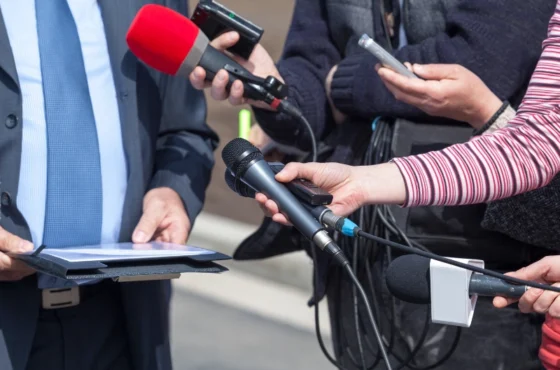 Group of journalists holding microphones, recorders, and smartphones towards a person in a suit during a press interview or media briefing outdoors.