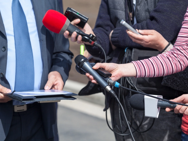 Group of journalists holding microphones, recorders, and smartphones towards a person in a suit during a press interview or media briefing outdoors.