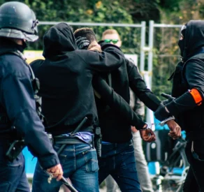 Heavily armed police officers detaining individuals in hooded clothing during a tense confrontation, with one person being handcuffed, suggesting a protest or civil unrest situation.