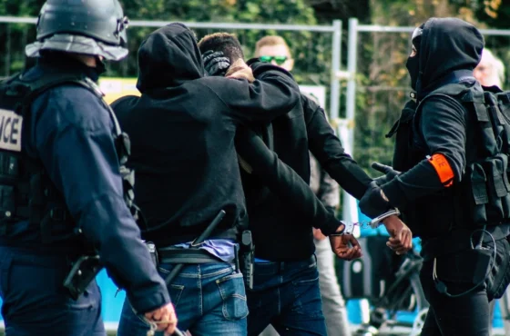 Joint Statement from The Decalogue Society of Lawyers 2 Heavily armed police officers detaining individuals in hooded clothing during a tense confrontation, with one person being handcuffed, suggesting a protest or civil unrest situation.