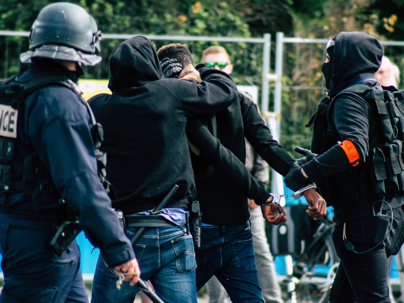 Heavily armed police officers detaining individuals in hooded clothing during a tense confrontation, with one person being handcuffed, suggesting a protest or civil unrest situation.