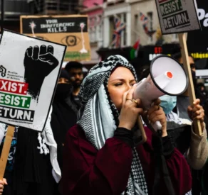 Pro-Palestinian protest with a woman in a keffiyeh using a megaphone, surrounded by demonstrators holding signs that read 'Exist. Resist. Return.' and 'Your indifference kills' during a public demonstration.