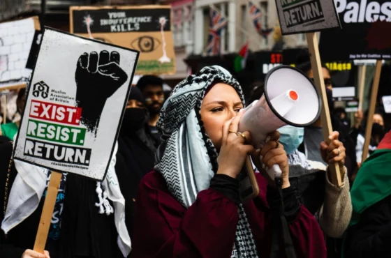 Condemning Targeted Protests 1 Pro-Palestinian protest with a woman in a keffiyeh using a megaphone, surrounded by demonstrators holding signs that read 'Exist. Resist. Return.' and 'Your indifference kills' during a public demonstration.