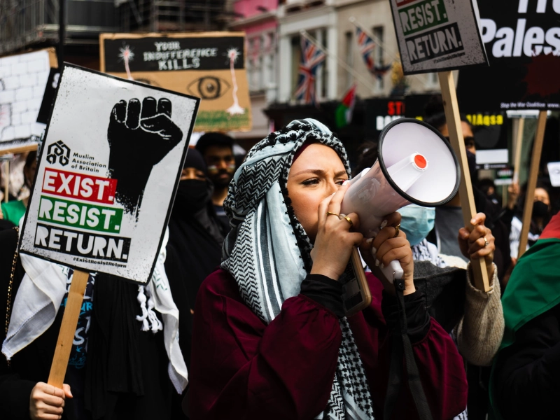 Pro-Palestinian protest with a woman in a keffiyeh using a megaphone, surrounded by demonstrators holding signs that read 'Exist. Resist. Return.' and 'Your indifference kills' during a public demonstration.